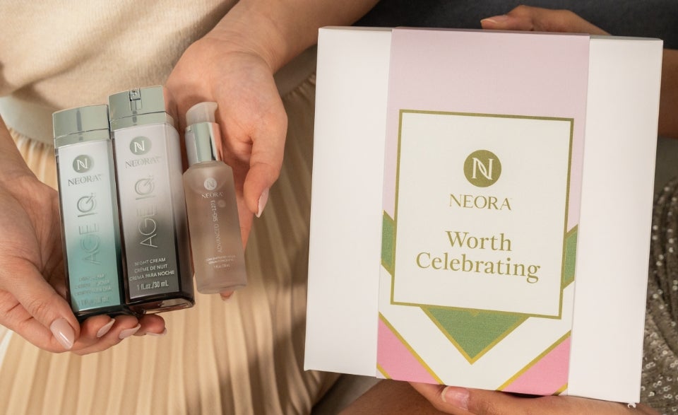 A woman holds Neora’s Age IQ Day and Night Creams bottles & Sig Serum in her hands next to a Neora branded box that says “Worth Celebrating.”