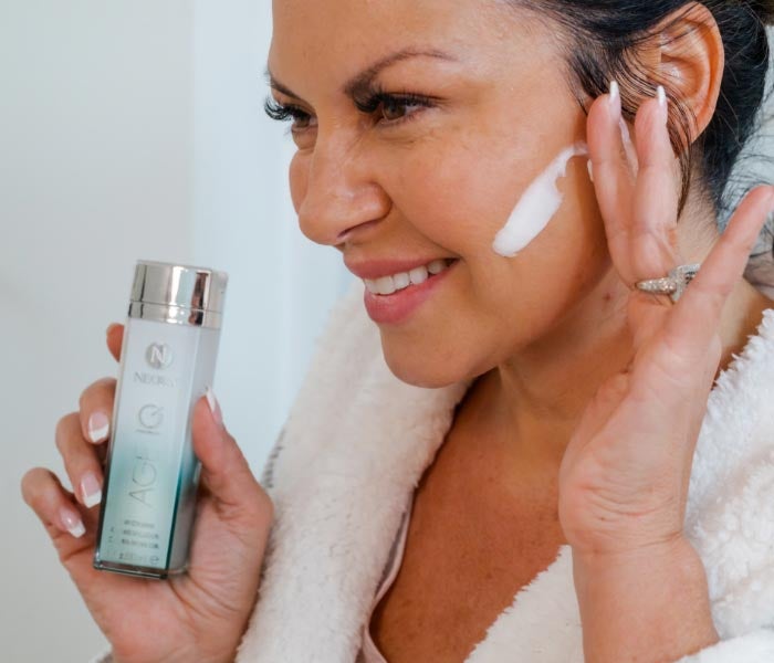 Photo of woman in bathrobe applying Neora Day Cream to her cheek.