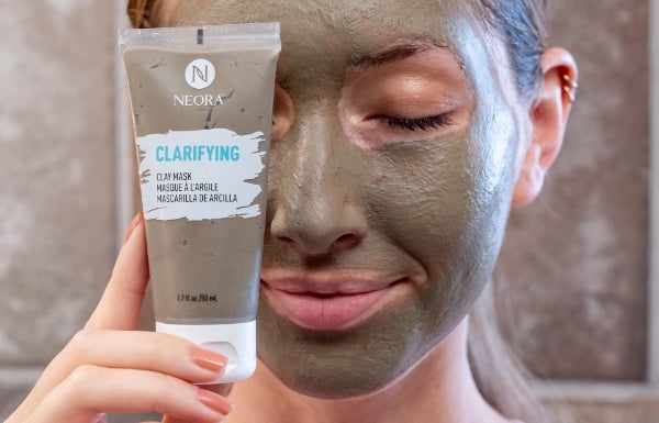 A close up of a woman’s face covered with Neora’s Clarifying Clay mask. She holds up a tube of this product in front of the right side of her face.